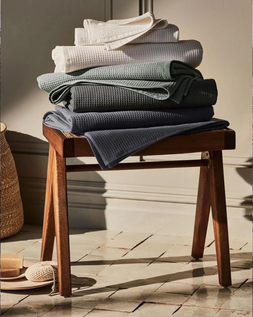 Stack of folded waffle-weave towels in neutral hues on a wooden stool, set against a neutral background. Home decor and textile.