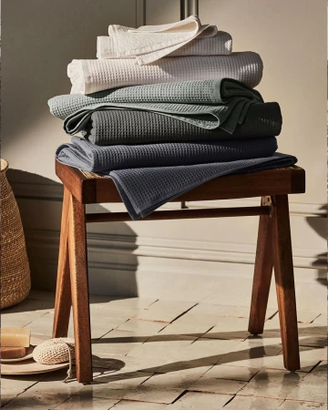 Stack of folded waffle-weave towels in neutral hues on a wooden stool, set against a neutral background. Home decor and textile.