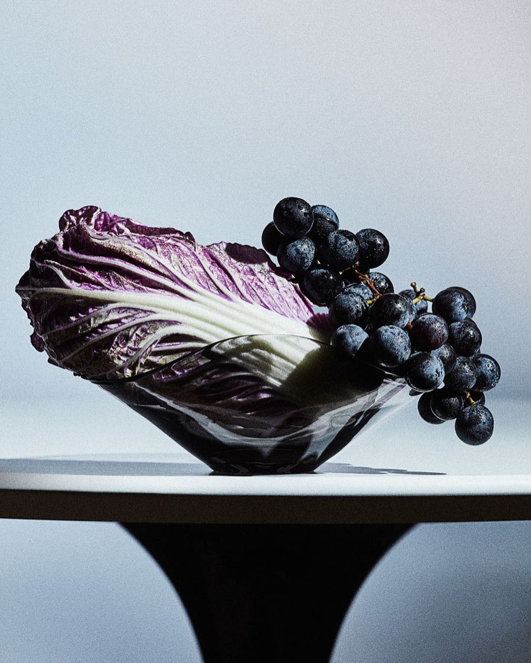 Still life photograph featuring a head of purple cabbage and a bunch of black grapes in a glass bowl, set against a gradient background.
