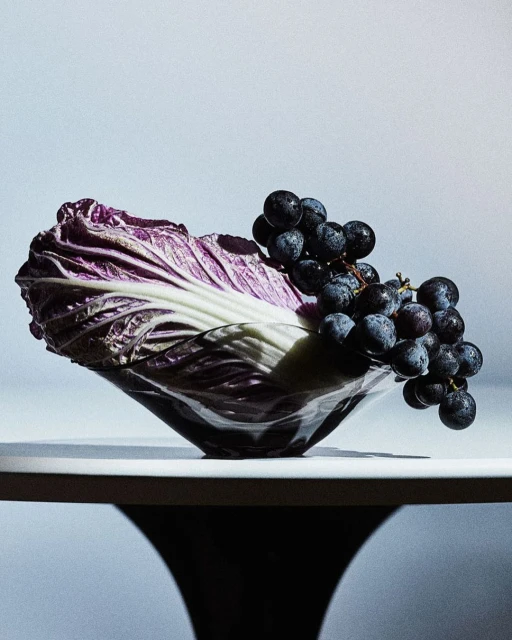 Still life photograph featuring a head of purple cabbage and a bunch of black grapes in a glass bowl, set against a gradient background.