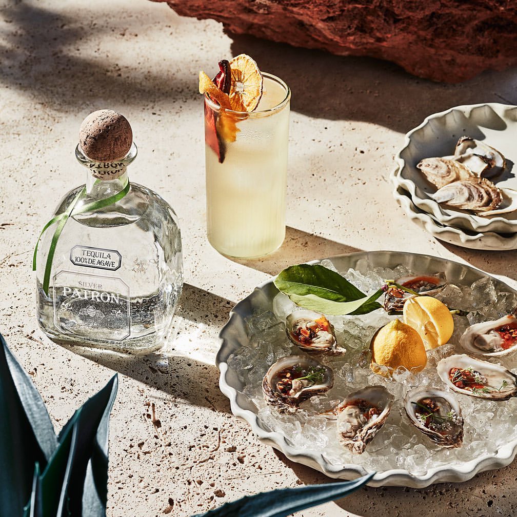 Tequila bottle and cocktail on table next to oysters on ice in a bright setting, suggesting a sophisticated food and beverage presentation.