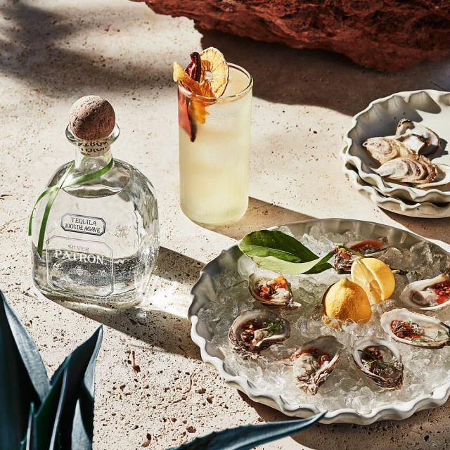 Tequila bottle and cocktail on table next to oysters on ice in a bright setting, suggesting a sophisticated food and beverage presentation.