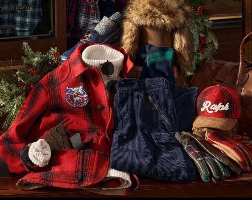 Red and black plaid coat, blue corduroy pants, and leather accessories are displayed with winter greenery for a festive, seasonal aesthetic.