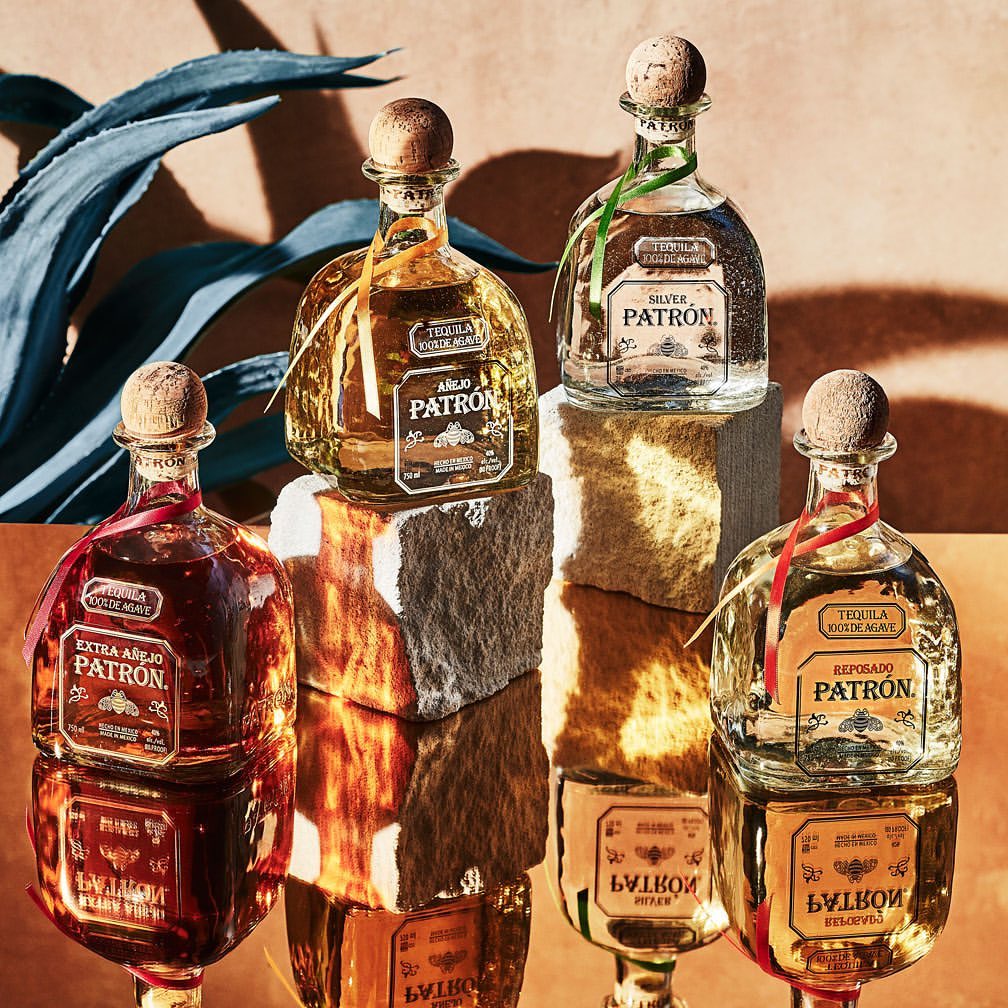 Five Patron tequila bottles arranged on mirrored surface, showing Extra Anejo, Anejo, Silver, and Reposado varieties with agave plant background.