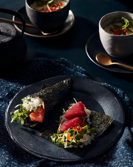 Close-up of two seaweed-wrapped temaki sushi rolls with tuna, salmon, rice, and greens served on a dark plate with soups in background.