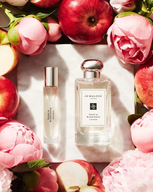 Two Jo Malone perfume bottles surrounded by pink peonies and sliced red apples on a white marble surface in bright, soft studio lighting.