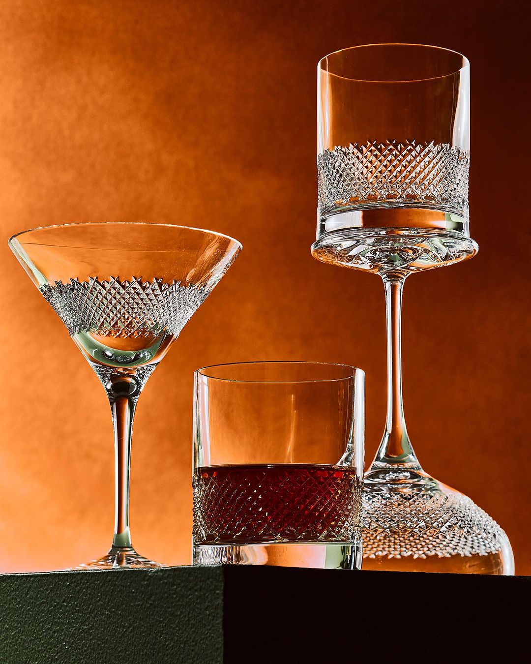 Still life of a martini glass, a lowball glass, and a tall glass with decorative patterns, set against an orange background.