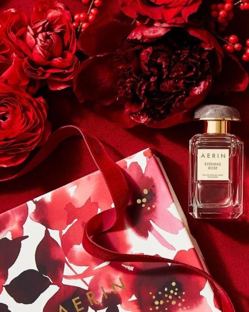 AERIN Evening Rose perfume bottle and gift box surrounded by deep red roses and a satin ribbon, set against a matching red fabric background.