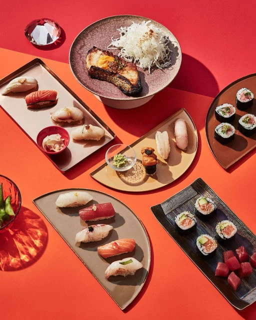 Overhead shot of vibrant assortment of sushi and Japanese cuisine on colorful trays and dishes against a red background.