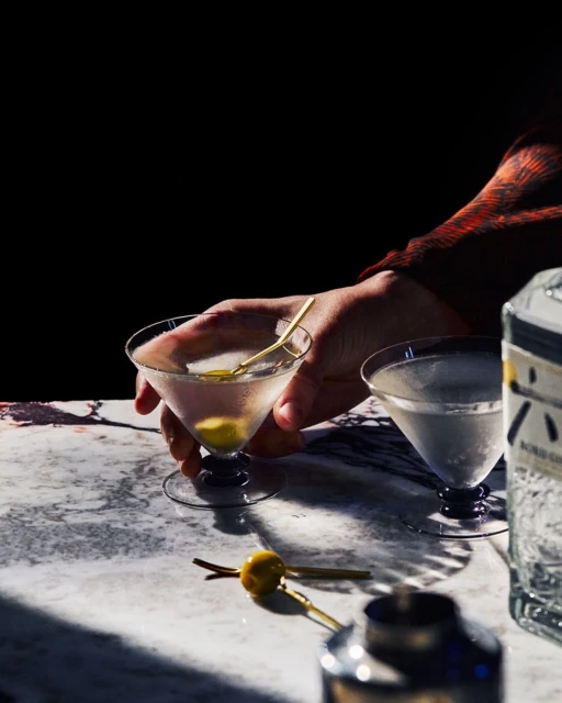 Hand holding a martini glass with an olive garnish, set on a marble table with a cocktail shaker and another martini in background.