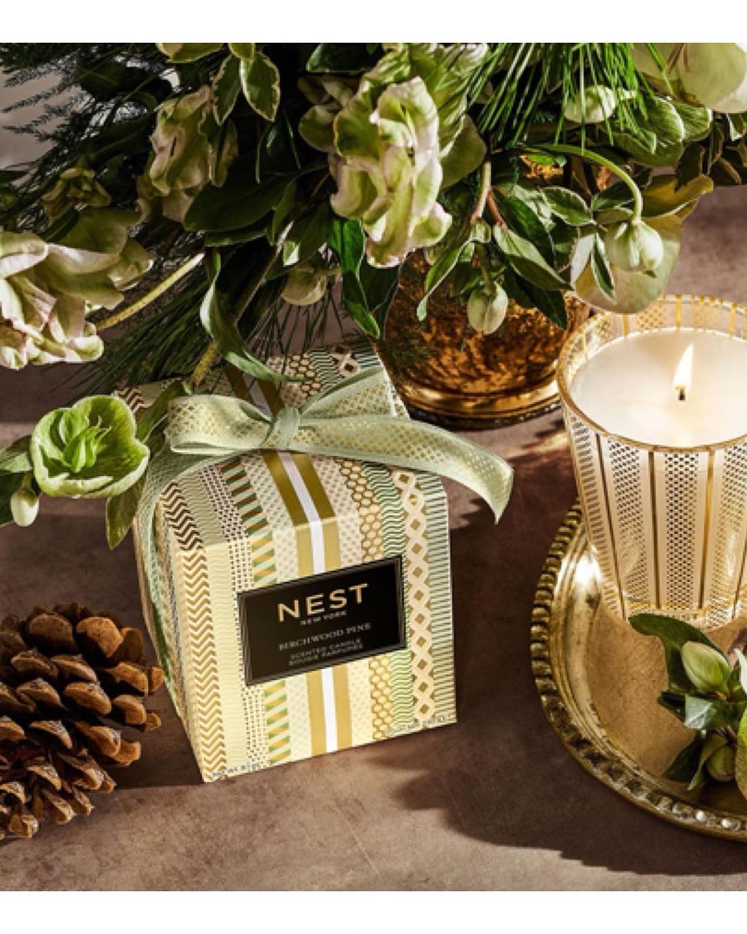 Elegant still life featuring a Nest Birchwood Pine candle, pinecone, and flower arrangement, set against a soft, neutral backdrop.