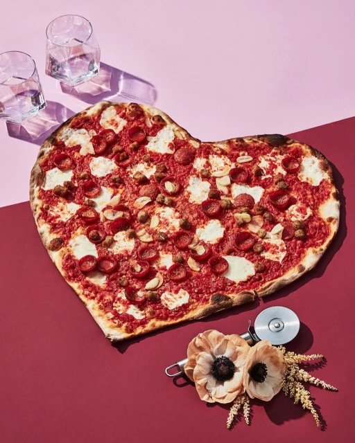 Overhead shot of a heart-shaped pizza on a split pink and burgundy background, alongside two glasses and decorative flowers. Food and beverage still life.
