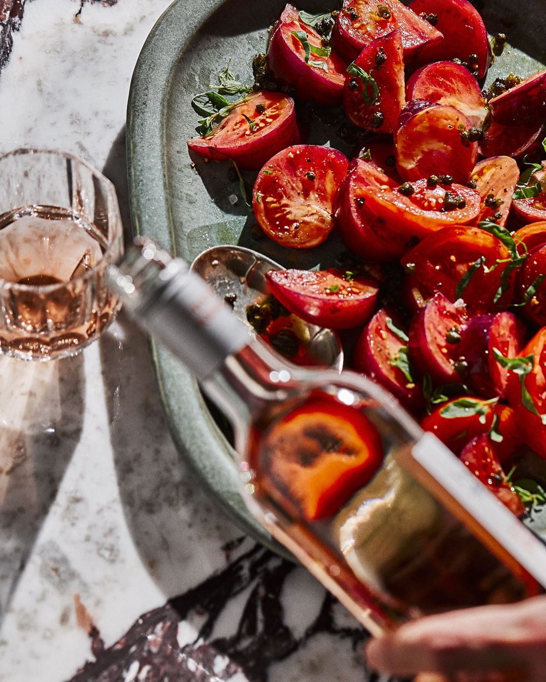 Overhead shot of a bottle of rosé wine being poured next to a dish of sliced tomatoes with fresh herbs and black peppercorns.