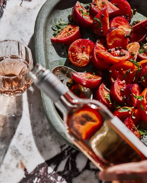 Overhead shot of a bottle of rosé wine being poured next to a dish of sliced tomatoes with fresh herbs and black peppercorns.