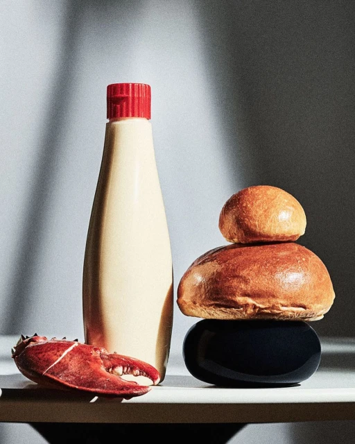 Still life shot of food ingredients: a bottle of sauce, a black bowl, buns stacked, and lobster claw, lit by natural light.