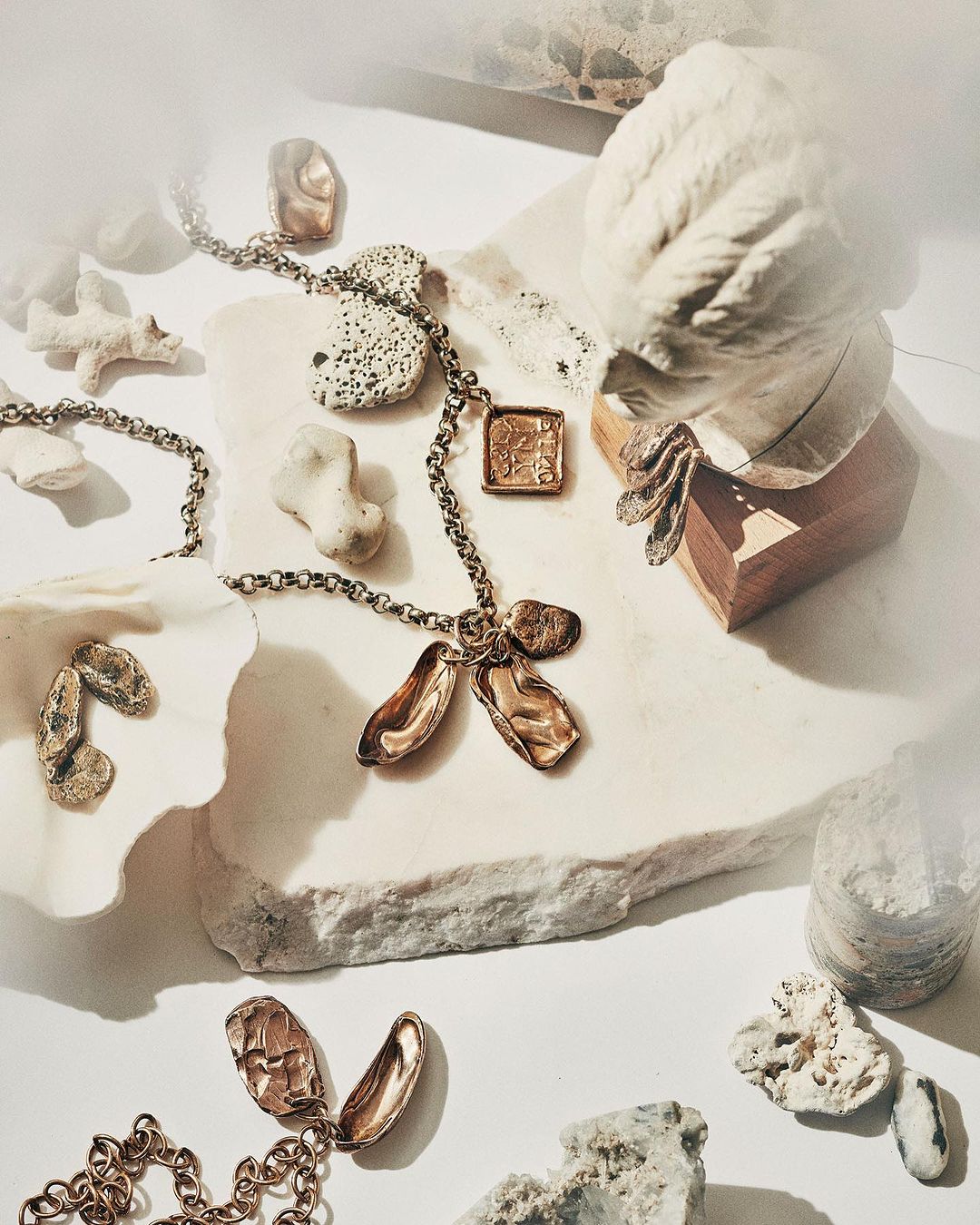 Necklace with bronze charms displayed on a white marble surface next to a bust and assorted stones and shells in a studio setting.
