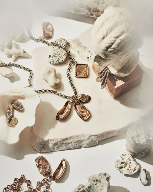 Necklace with bronze charms displayed on a white marble surface next to a bust and assorted stones and shells in a studio setting.