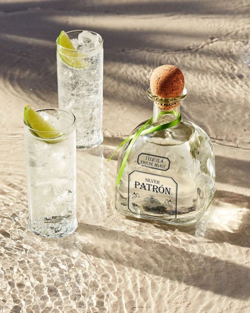 Bottle of Patron tequila with two cocktails on a wet sandy surface, suggesting a beach setting with drink photography.