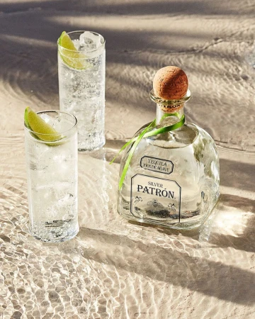 Bottle of Patron tequila with two cocktails on a wet sandy surface, suggesting a beach setting with drink photography.