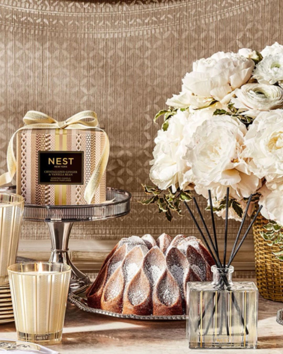 Still life of a NEST New York candle and room diffuser with a bundt cake, and white flower arrangement on a table.