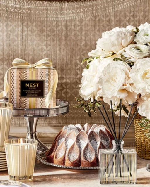 Still life of a NEST New York candle and room diffuser with a bundt cake, and white flower arrangement on a table.