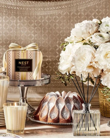 Still life of a NEST New York candle and room diffuser with a bundt cake, and white flower arrangement on a table.
