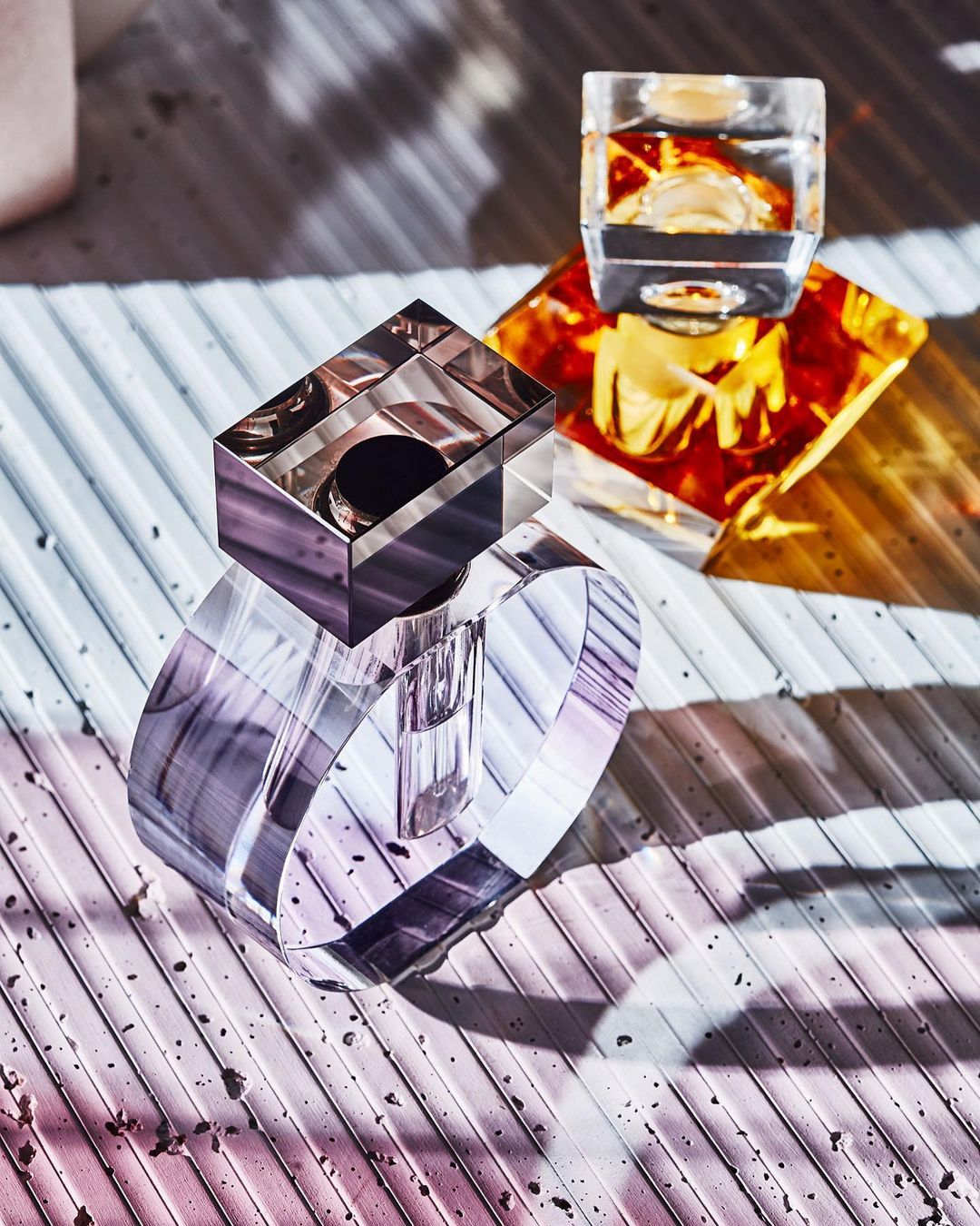 Geometric crystal perfume bottles with lilac and amber color gradients, displayed on a ribbed surface with strong light and shadow.