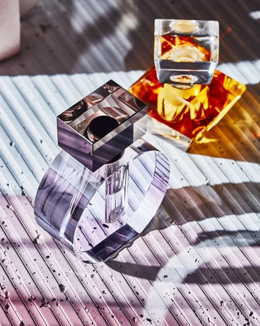 Geometric crystal perfume bottles with lilac and amber color gradients, displayed on a ribbed surface with strong light and shadow.