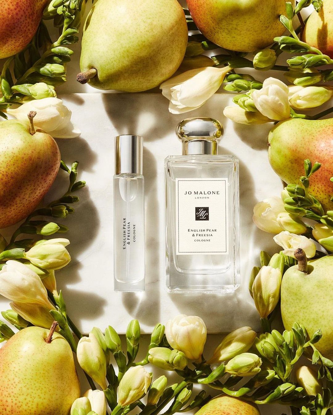 Two bottles of Jo Malone London cologne surrounded by green pears, white freesia flowers, and leaves on a white marble surface.