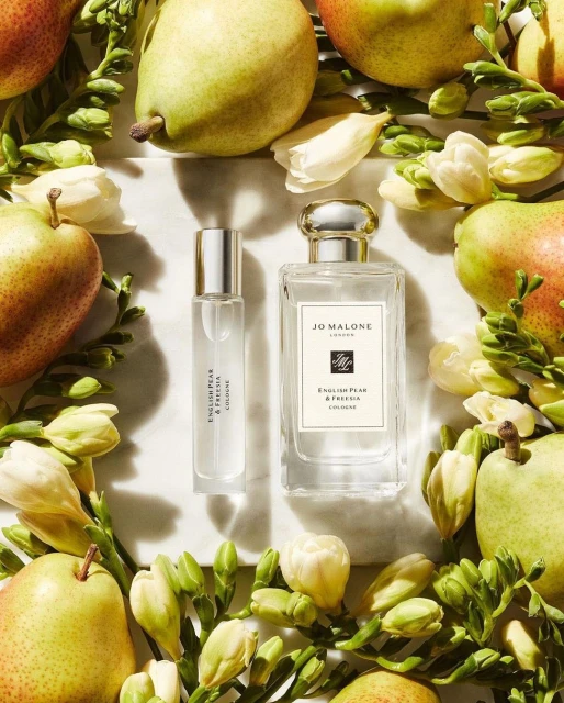 Two bottles of Jo Malone London cologne surrounded by green pears, white freesia flowers, and leaves on a white marble surface.