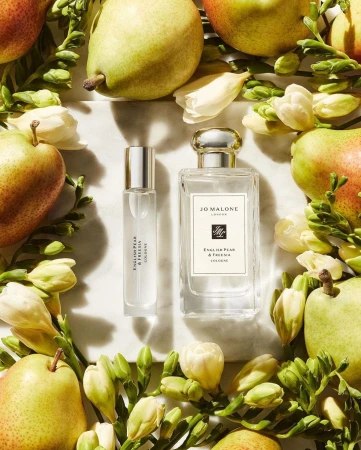 Two bottles of Jo Malone London cologne surrounded by green pears, white freesia flowers, and leaves on a white marble surface.
