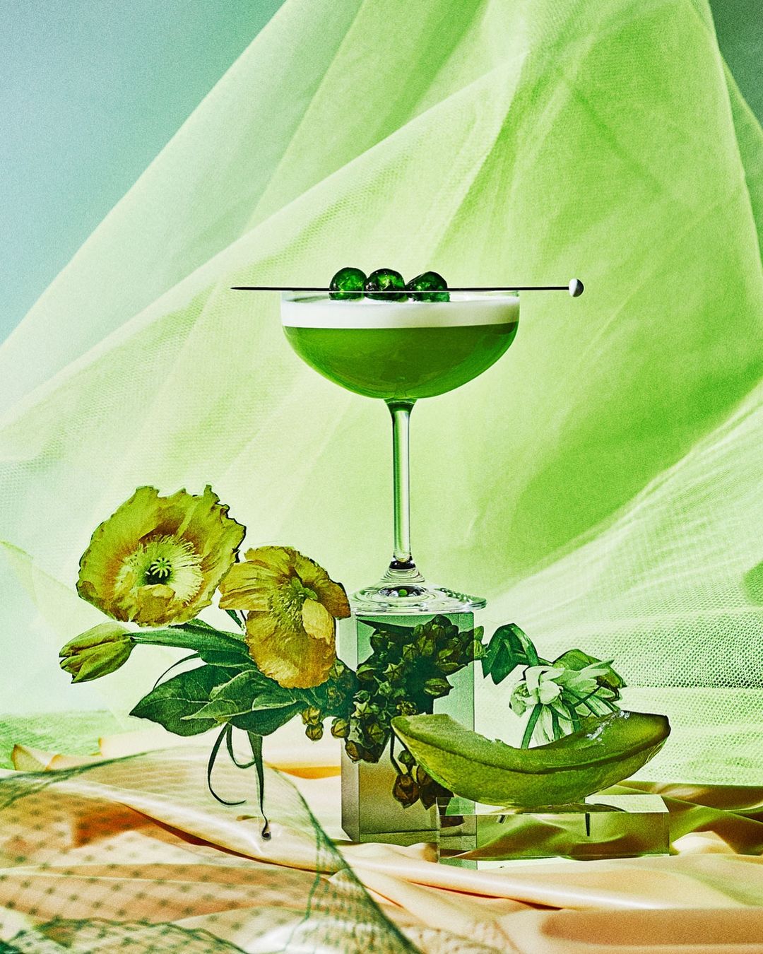 Green cocktail in a glass with fruit garnish and flowers on a decorative table, set against a light green background with sheer fabric.