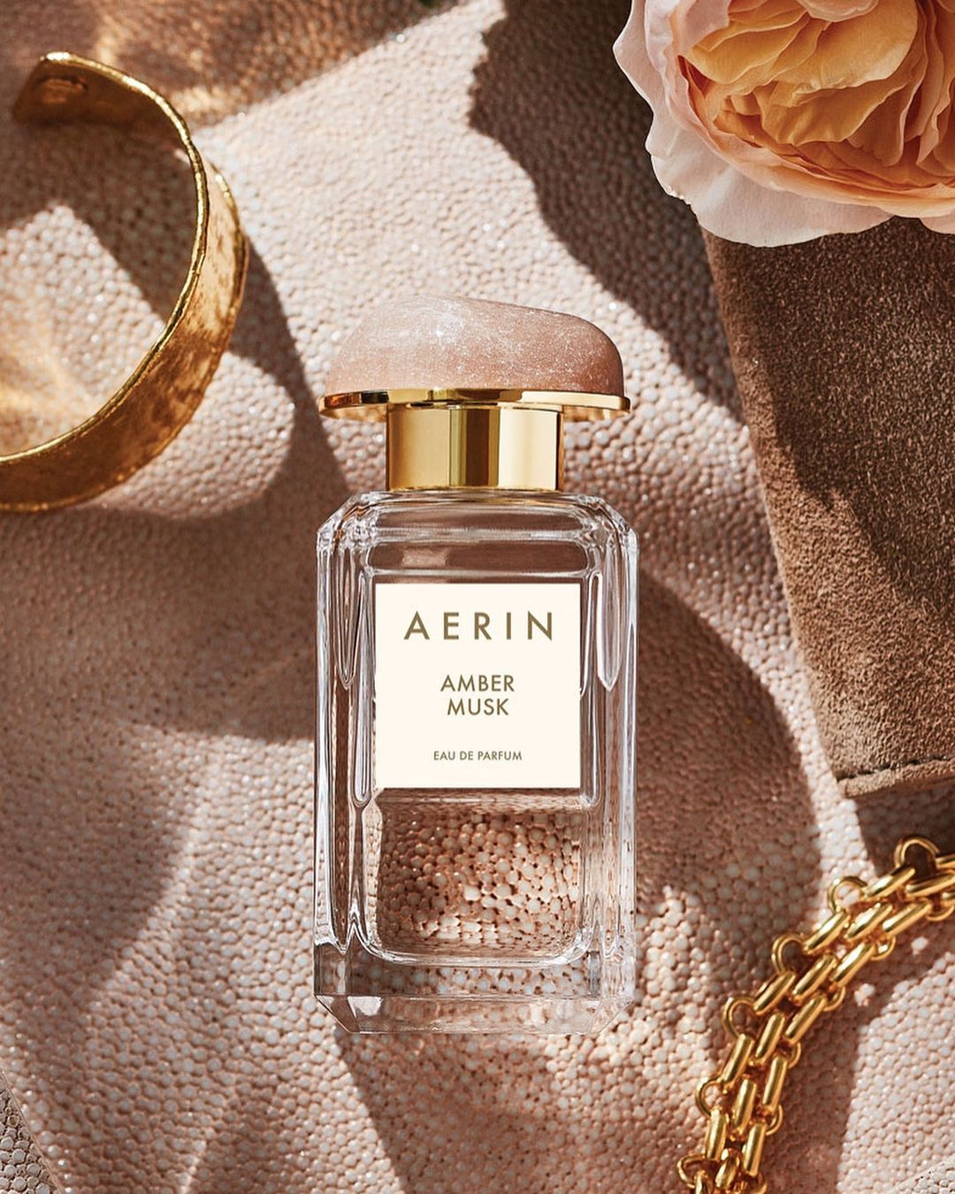 Close-up of Aerin Amber Musk perfume bottle with gold accents on a textured surface, complemented by a peach-colored rose and gold bangle.