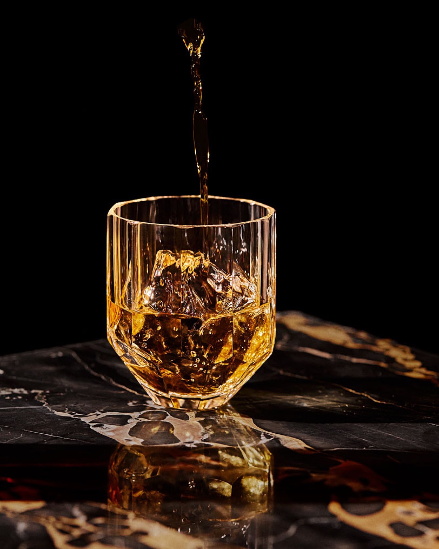 Amber liquid pouring into a faceted crystal glass with ice cubes on a dark marble surface.