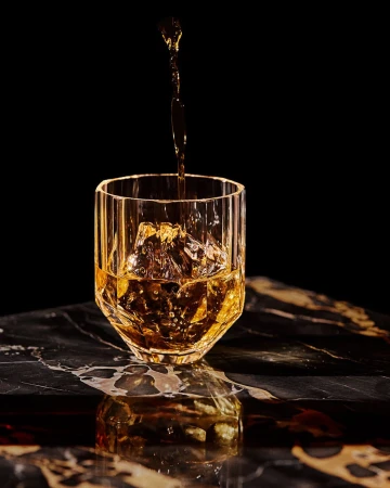 Amber liquid pouring into a faceted crystal glass with ice cubes on a dark marble surface.