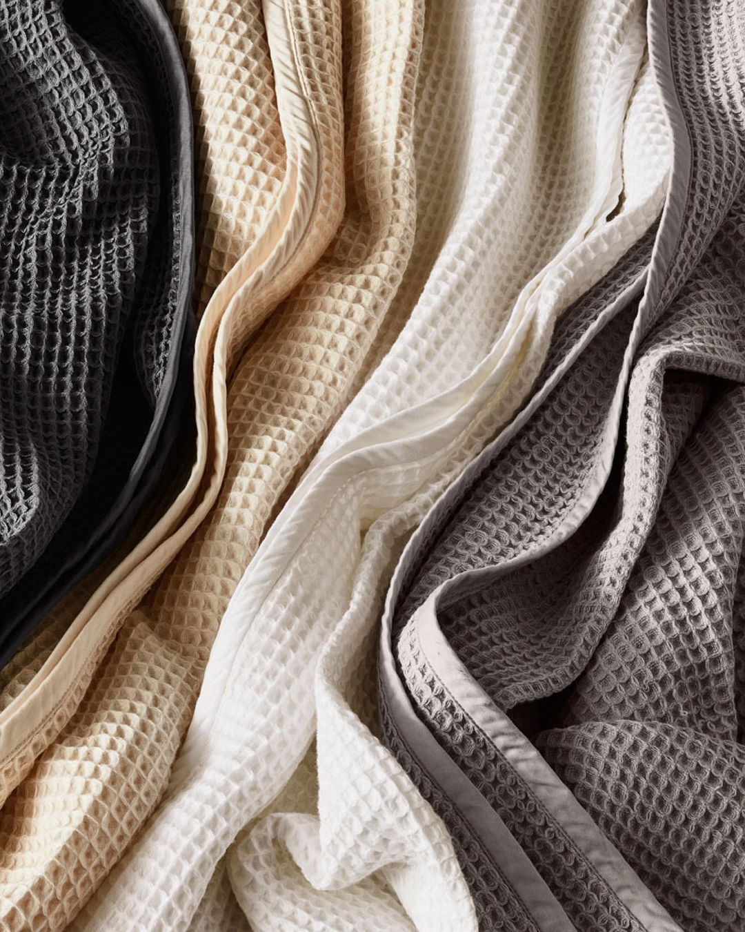 Arrangement of four textured waffle blankets in a high-angle shot, displaying a range of neutral colors from dark grey to beige to white.