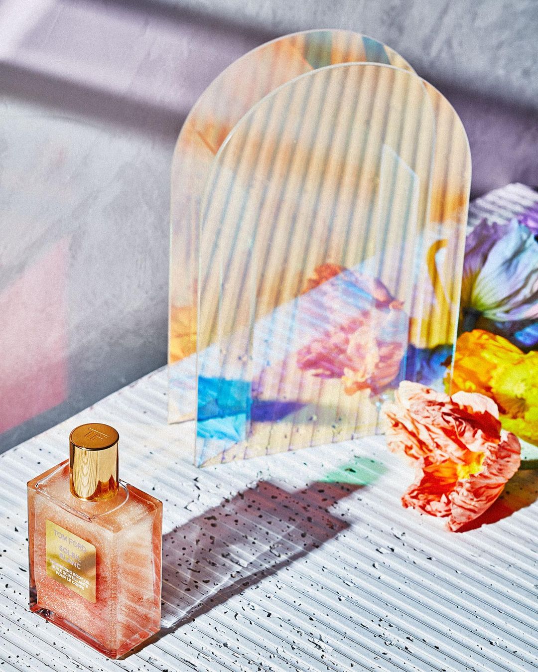 Close-up of a perfume bottle with a golden cap, flowers, and transparent arch, set against a textured white surface in a studio setting.