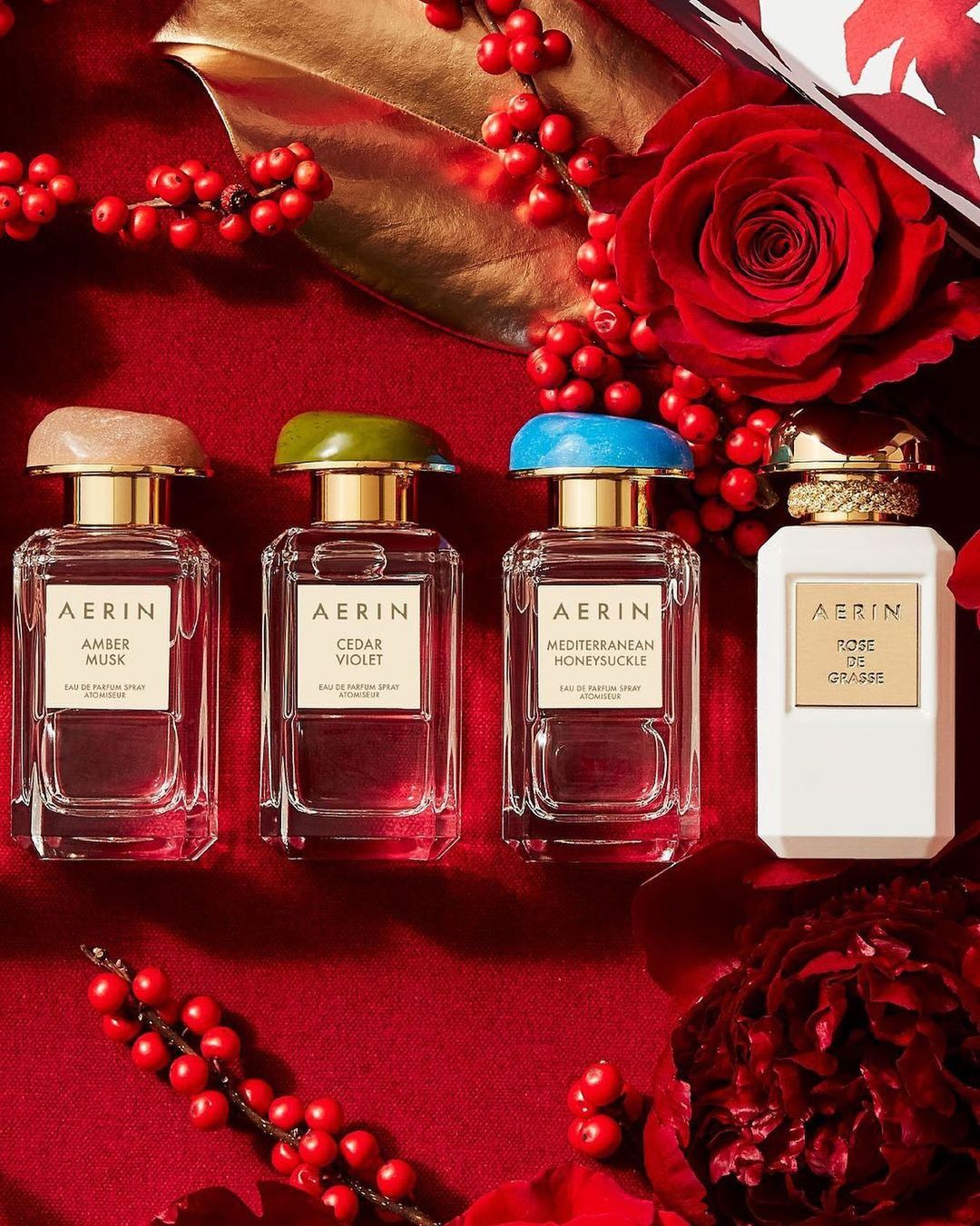 Four AERIN perfume bottles are displayed against a red background with floral and berry accents, showcasing different colored caps.