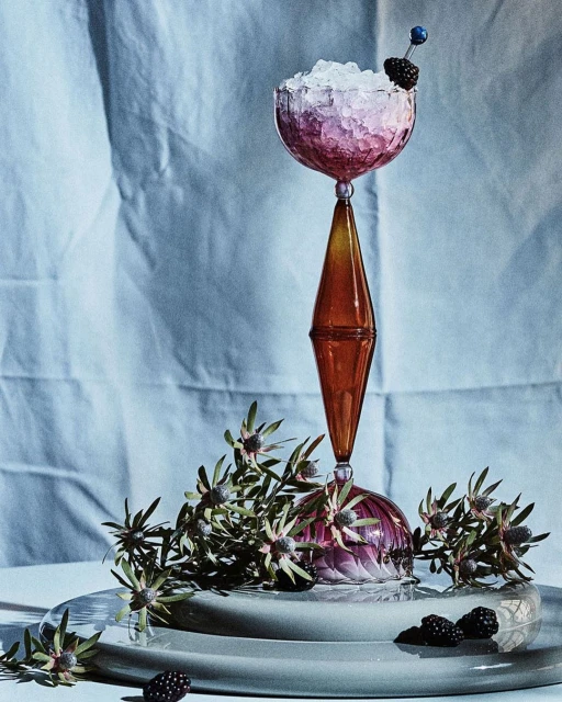 Elegant stemmed glass filled with crushed ice and a blackberry garnish, set on a plate next to greenery and scattered blackberries.