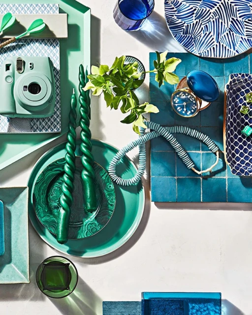 A flat lay photo features various objects including a mint green camera, twisted green candles, a blue tile, and tableware arranged on a white surface.