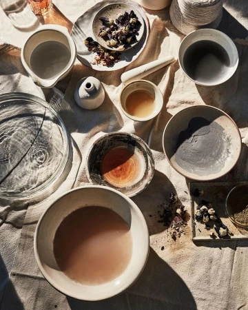 Overhead shot featuring multiple ceramic bowls containing liquids and powders, arranged on a textured beige cloth surface with soft shadows.