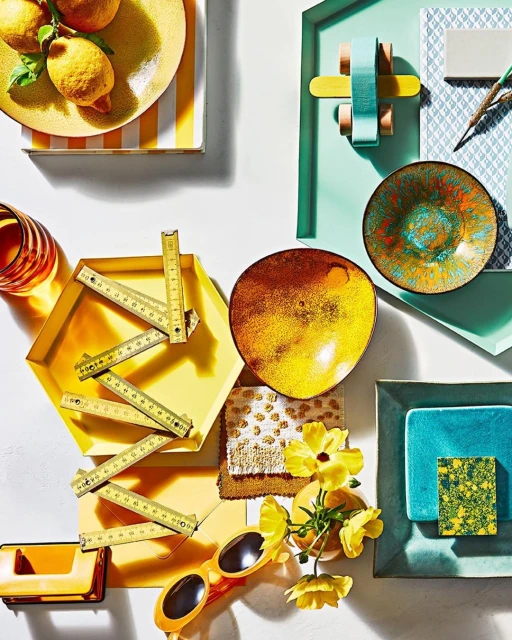 Overhead shot of an arrangement of colorful objects including lemons, plates, flowers, sunglasses, and a folding ruler on a white surface.