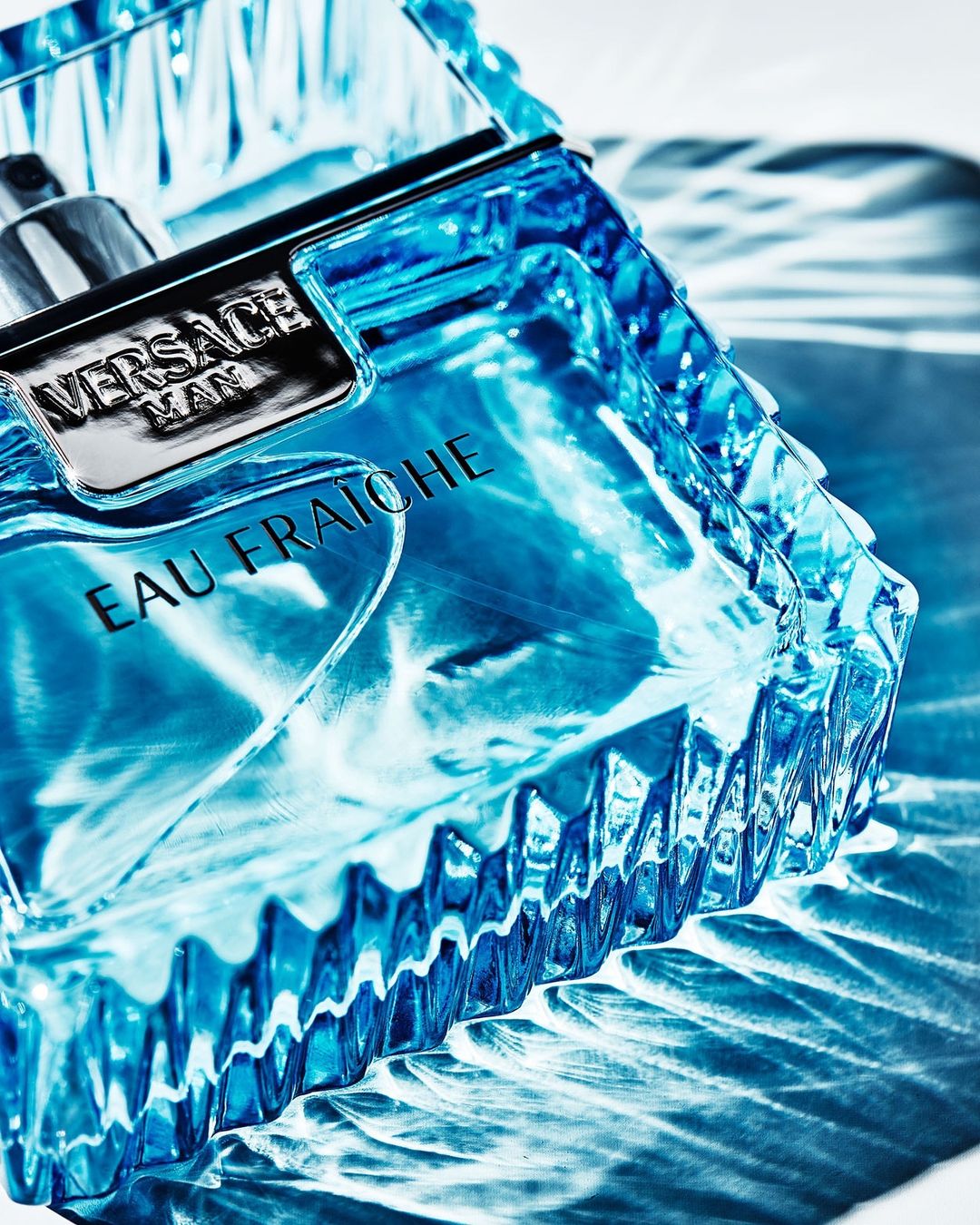 Close-up studio shot of Versace Man Eau Fraiche cologne bottle with a textured blue glass design and silver accents against a white background.