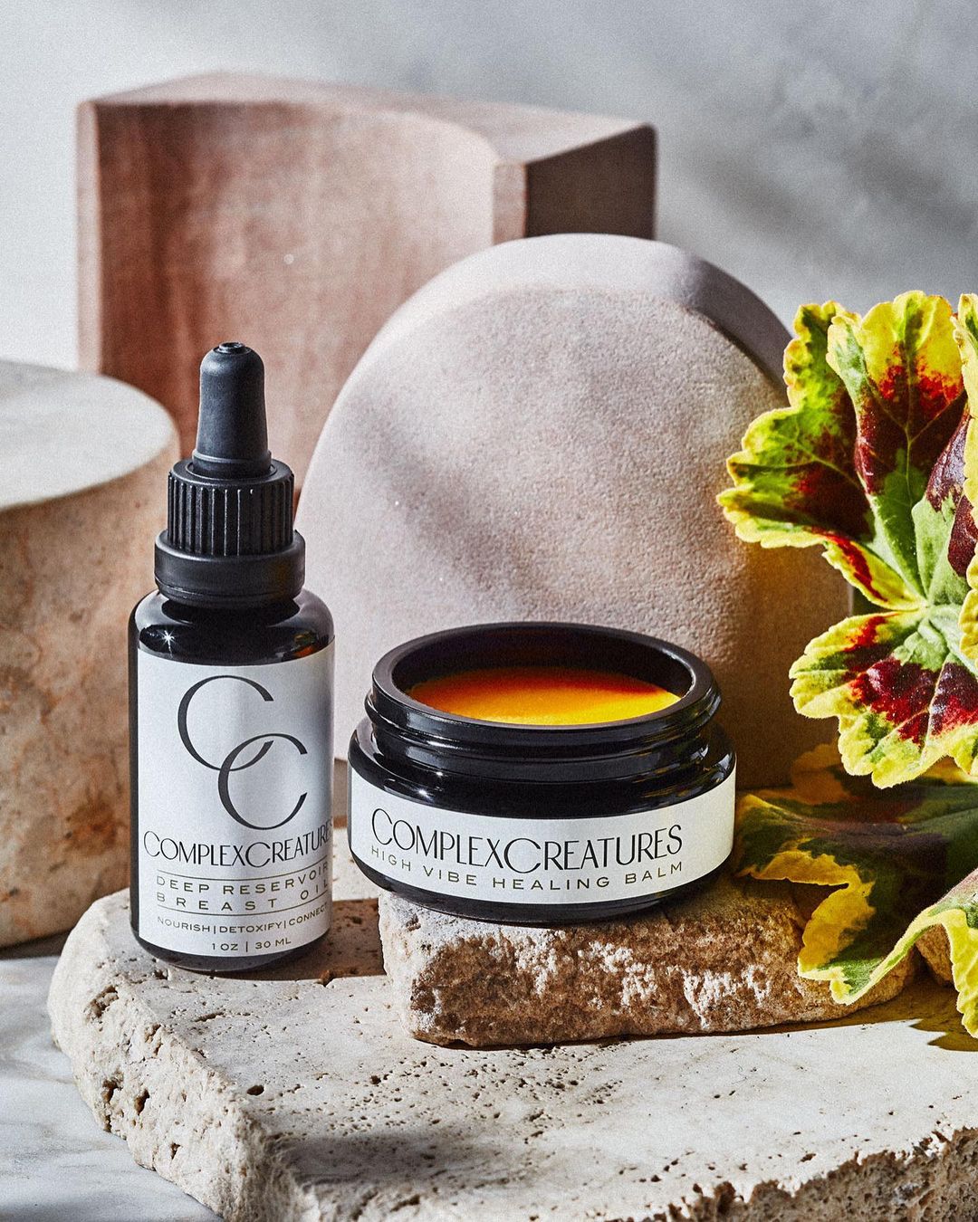 Complex Creatures skincare products: Deep Restorative Heart Oil and High Vibe Healing Balm, presented on a textured stone surface.