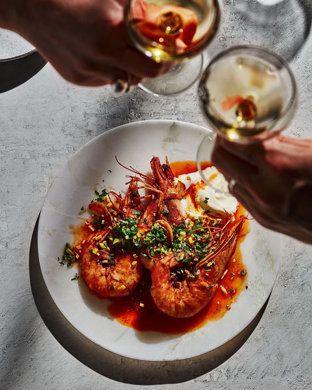 Overhead shot of shrimp dish on marble plate, with wine glasses held by two people. Food and beverage photography with bright lighting.