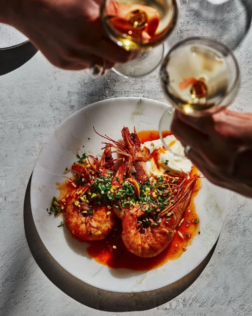 Overhead shot of shrimp dish on marble plate, with wine glasses held by two people. Food and beverage photography with bright lighting.