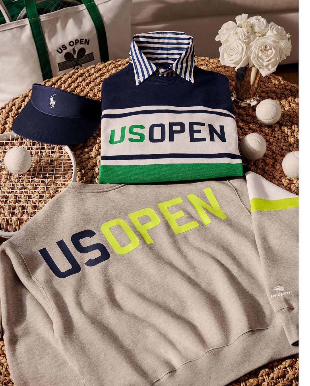 Still life of tennis-themed Ralph Lauren clothing and accessories, including shirts, sweater, tote bag, visor, tennis balls, and racket.