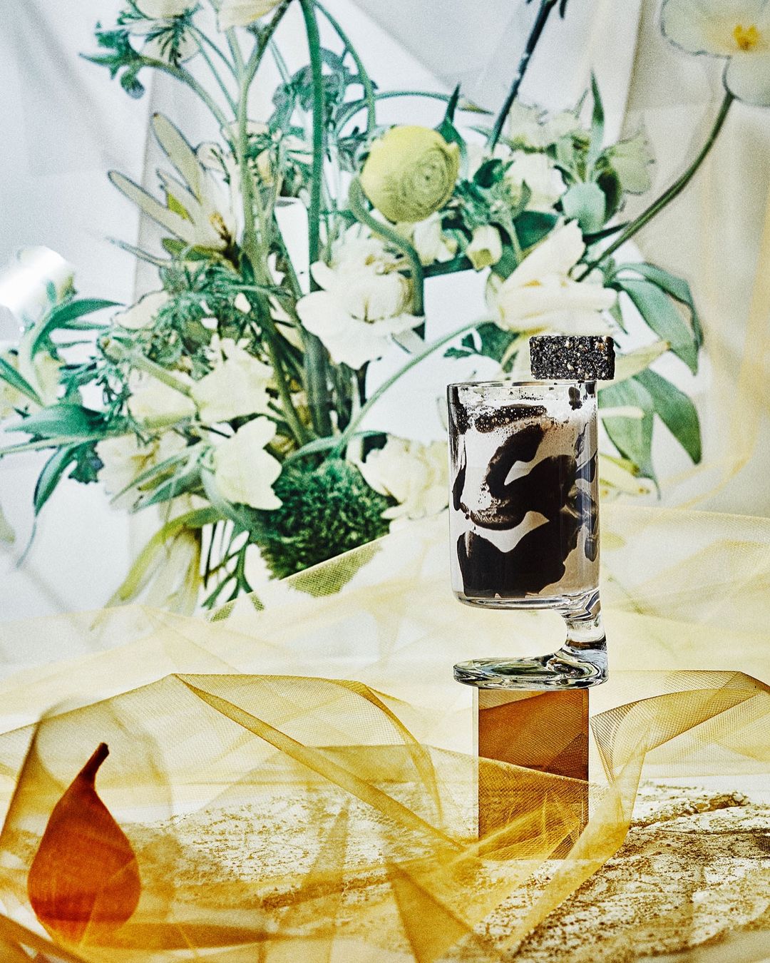 Still life photo of a modern glass vase with black and white brushstrokes filled with white liquid, flowers in the background.