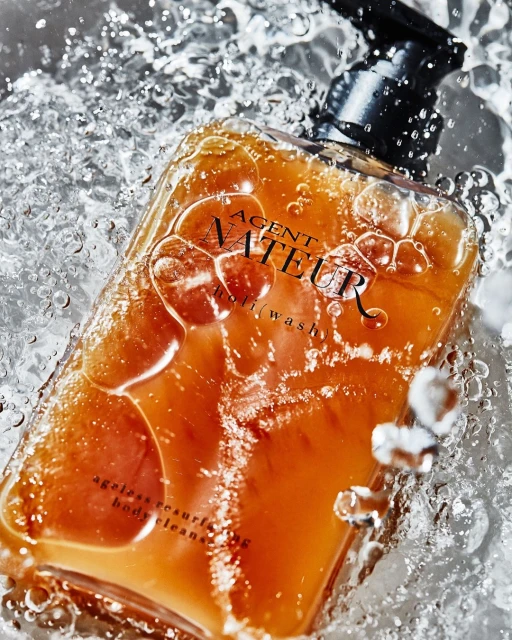 Agent Nateur body wash product shot, clear bottle with amber liquid, surrounded by water droplets and splashes on a neutral background.
