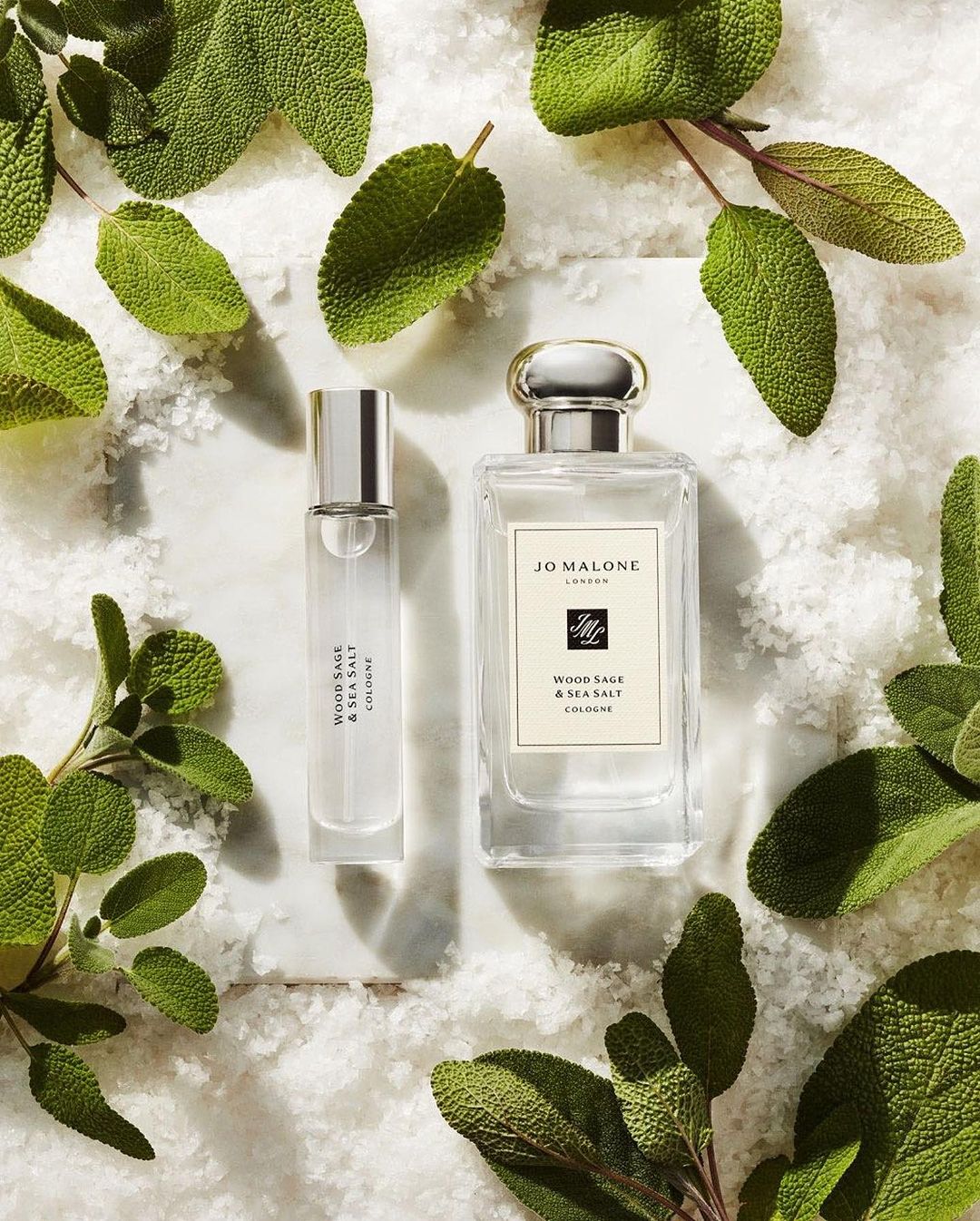 Two Jo Malone Wood Sage & Sea Salt cologne bottles on a marble surface, surrounded by fresh sage leaves and scattered salt granules.